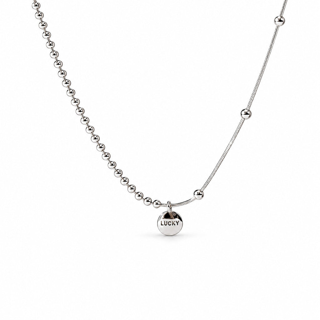 Sterling Silver Eira necklace with round LUCKY medallion, beaded and smooth chain sections, in the existing cropped tabletop view with white studio treatment and soft shadow; AI studio edit and enhancement of the existing supplier source view; chain ends extend beyond the frame.