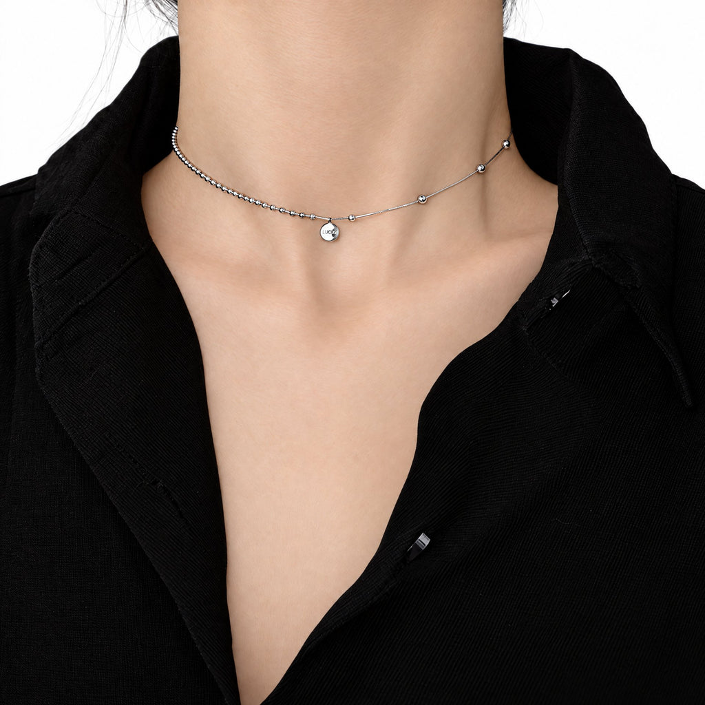 Sterling Silver Eira necklace with a small round LUCKY medallion in the existing worn neckline crop on a black collared shirt, set against white studio surroundings; AI studio edit and enhancement of the existing supplier source view; only visible chain sections are shown.