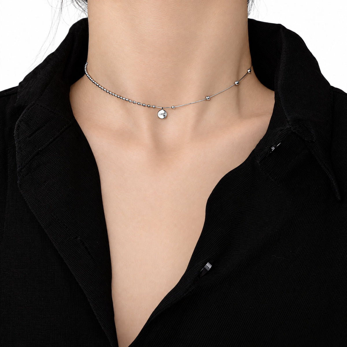 Sterling Silver Eira necklace with a small round LUCKY medallion in the existing worn neckline crop on a black collared shirt, set against white studio surroundings; AI studio edit and enhancement of the existing supplier source view; only visible chain sections are shown.